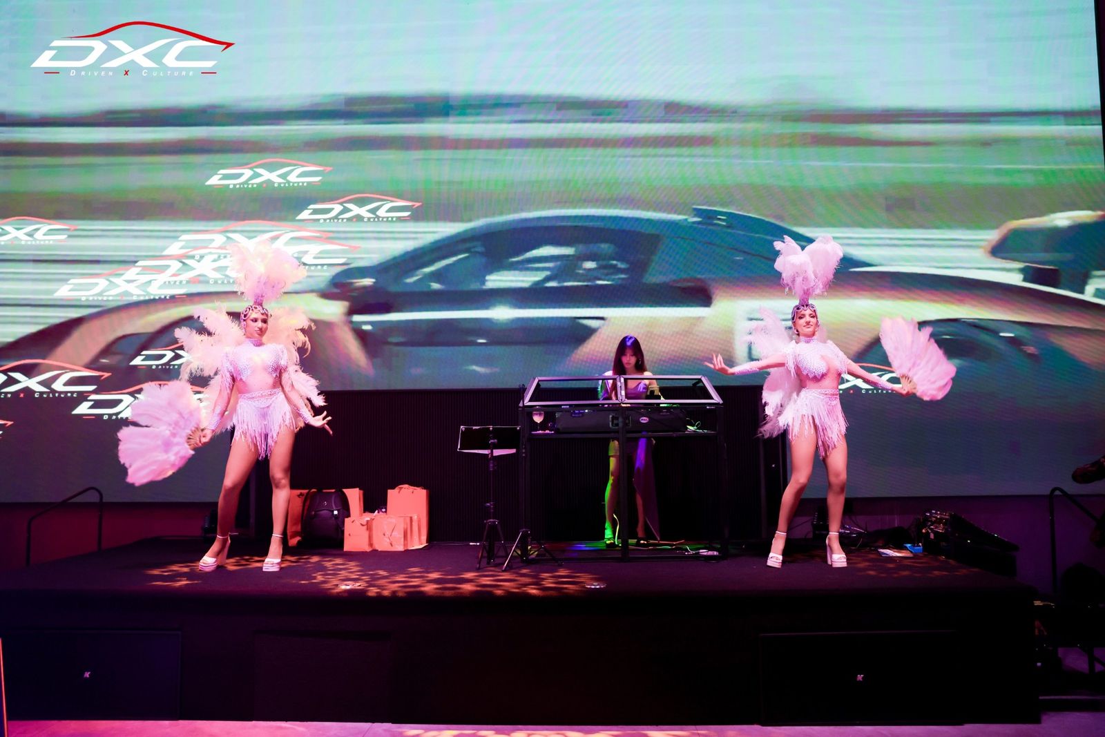 A DJ and two showgirl dancers in white feathers on stage in front of the DXC screen